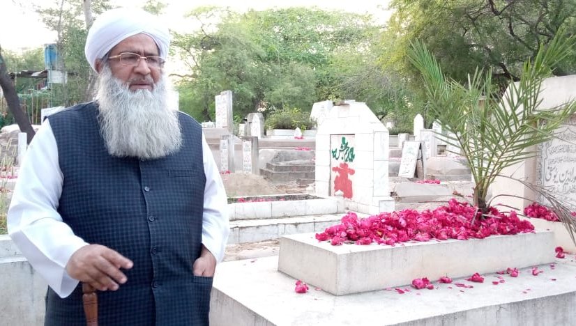 Shaykh Muhammad Mohsin Munawar Yousafi visiting a spiritual grave (Mazaar) or graveyard, standing next to a tomb decorated with pink flowers, holding a cane.