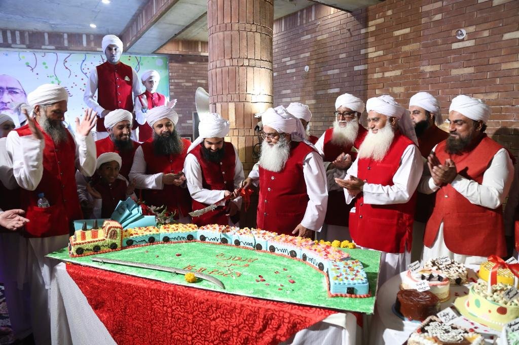 Shaykh Muhammad Mohsin Munawar Yousafi and a group of senior scholars wearing matching red vests, cutting a large, custom-designed cake shaped like a train during an institutional celebration or birthday event.
